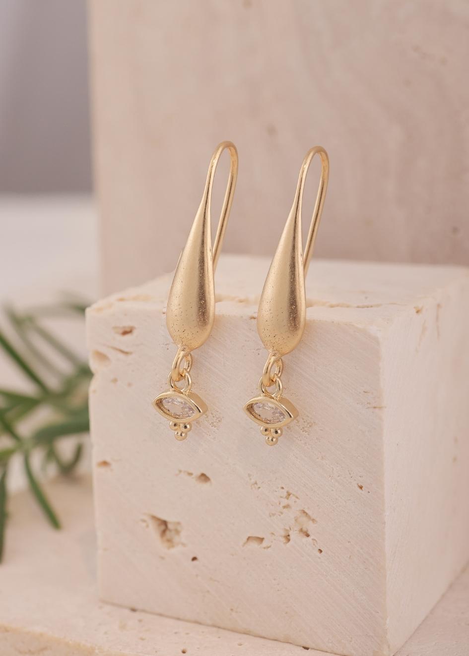 עגילי שנהב זרקון שקוף של הדס קוקה – עגילי ציפוי זהב עדינים עם תליון בצורת עין משובצת זרקון שקוף, מצולמים על רקע אבן טבעית.